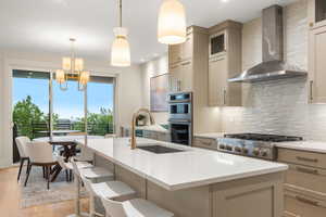 Kitchen with decorative backsplash, wall chimney exhaust hood, light countertops, a chandelier, and a kitchen island with sink