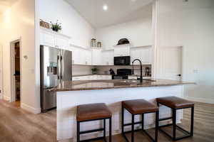 Kitchen with black appliances, high vaulted ceiling, a kitchen breakfast bar, white cabinetry, and an island with sink