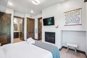 Bedroom featuring wood finished floors, a glass covered fireplace, ensuite bath, and recessed lighting