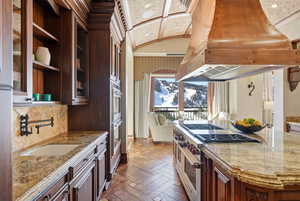 Kitchen featuring custom exhaust hood, dark brown cabinets, dark stone counters, open shelves, and stainless steel appliances