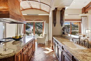 Kitchen with light stone counters, premium range hood, brown cabinetry, recessed lighting, and lofted ceiling