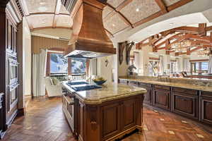 Kitchen with dark brown cabinetry, lofted ceiling, light stone countertops, custom exhaust hood, and appliances with stainless steel finishes