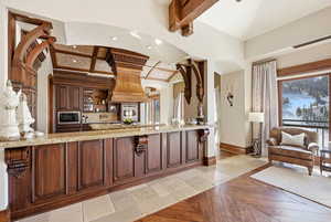 Kitchen featuring light stone countertops, beam ceiling, stainless steel microwave, stone tile flooring, and dark brown cabinets