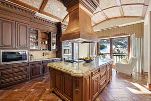 Kitchen with light stone counters, custom exhaust hood, glass insert cabinets, stainless steel appliances, and lofted ceiling