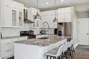 Kitchen with glass insert cabinets, light stone countertops, wall chimney range hood, white cabinetry, and stainless steel appliances
