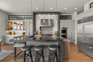 Kitchen with appliances with stainless steel finishes, light wood-style floors, exhaust hood, an island with sink, and tasteful backsplash