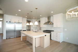 Kitchen featuring high quality appliances, wall chimney range hood, light wood-style flooring, white cabinetry, and a center island with sink