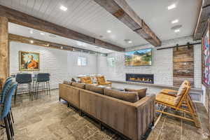 Living room with a barn door, beamed ceiling, a fireplace, brick wall, and stone finish floors