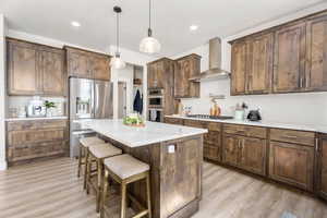Kitchen featuring stainless steel appliances, wall chimney exhaust hood, a kitchen breakfast bar, a center island, and light wood-type flooring