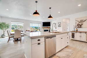 Kitchen featuring dishwasher, light wood finished floors, a fireplace, a center island with sink, and light countertops