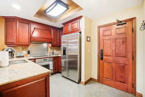 Kitchen featuring stainless steel appliances, decorative backsplash, light stone countertops, custom exhaust hood, and reddish brown cabinets