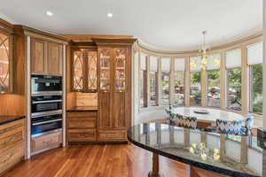 Kitchen with brown cabinetry, dark wood-style flooring, dark stone counters, a chandelier, and stainless steel double oven