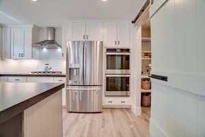 Kitchen featuring appliances with stainless steel finishes, dark countertops, wall chimney range hood, light wood finished floors, and white cabinetry