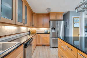 Kitchen featuring appliances with stainless steel finishes, light wood finished floors, decorative backsplash, dark stone counters, and glass insert cabinets