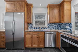 Kitchen featuring appliances with stainless steel finishes, backsplash, light stone countertops, brown cabinetry, and dark tile patterned floors