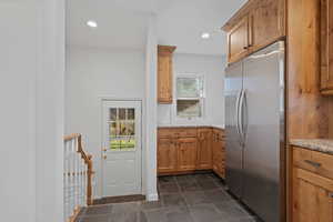 Kitchen with stainless steel refrigerator, recessed lighting, brown cabinets, light stone countertops, and dark tile patterned floors
