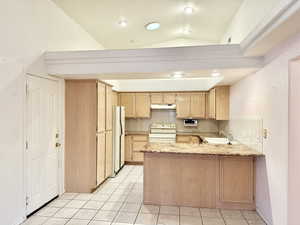 Kitchen with light brown cabinetry, a peninsula, light stone counters, white appliances, and lofted ceiling