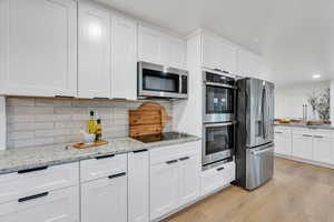 Kitchen with appliances with stainless steel finishes, light wood-style flooring, white cabinetry, decorative backsplash, and light stone countertops