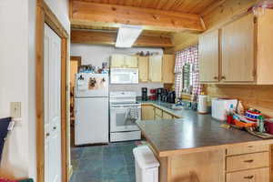 Kitchen with white appliances, a wooden ceiling with exposed beams, dark countertops, light brown cabinetry, and a peninsula