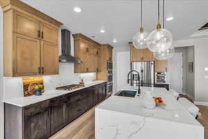 Kitchen with light wood-style floors, stainless steel appliances, wall chimney range hood, light stone counters, and hanging light fixtures