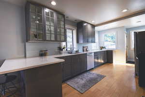 Kitchen featuring light wood-style floors, glass insert cabinets, stainless steel dishwasher, recessed lighting, and a breakfast bar area