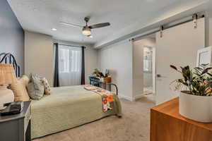 Bedroom featuring a textured ceiling, carpet flooring, a barn door, ensuite bath, and a ceiling fan