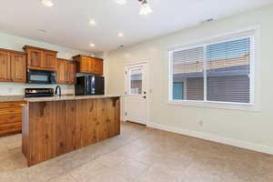 Kitchen with black appliances, brown cabinets, a kitchen island with sink, light stone countertops, and recessed lighting