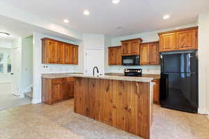 Kitchen featuring black appliances, brown cabinets, recessed lighting, light stone countertops, and an island with sink