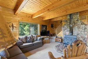 Living room with wood walls, a wood stove, a wood ceiling with exposed beams, carpet floors, and stairs