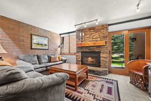 Living area with rail lighting, light tile patterned floors, a textured ceiling, a stone fireplace, and brick wall