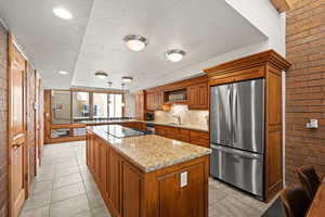 Kitchen featuring brown cabinetry, stainless steel fridge, a textured ceiling, a center island, and light stone countertops