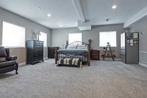 Carpeted bedroom with freestanding refrigerator, beam ceiling, multiple windows, and recessed lighting