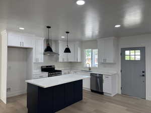 Kitchen with light wood finished floors, appliances with stainless steel finishes, wall chimney range hood, white cabinets, and recessed lighting