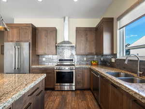 Kitchen with stainless steel appliances, wall chimney range hood, dark wood-style flooring, light stone countertops, and tasteful backsplash