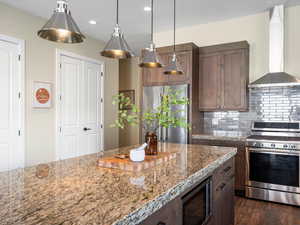 Kitchen with wall chimney range hood, appliances with stainless steel finishes, decorative backsplash, light stone countertops, and hanging light fixtures