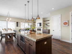 Kitchen featuring a glass covered fireplace, dark wood finished floors, dark brown cabinetry, stainless steel microwave, and decorative light fixtures