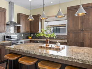 Kitchen with backsplash, wall chimney range hood, a kitchen bar, stainless steel gas range, and light stone counters