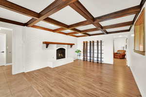 Unfurnished living room featuring light wood finished floors, beam ceiling, a fireplace, coffered ceiling, and brick wall