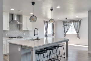 Kitchen featuring decorative backsplash, a kitchen island with sink, wall chimney exhaust hood, light wood-style floors, and a kitchen breakfast bar