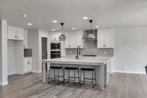 Kitchen featuring tasteful backsplash, light countertops, wall chimney range hood, a kitchen island with sink, and white cabinetry