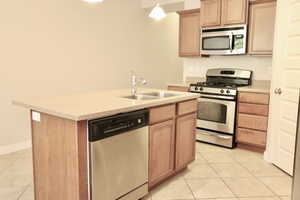 Kitchen with appliances with stainless steel finishes, light tile floors, light countertops, a center island with sink, and light brown cabinetry