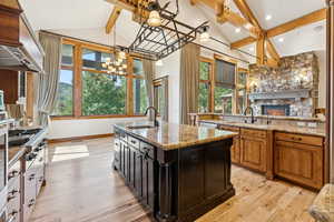 Kitchen with beamed ceiling, custom exhaust hood, light wood-type flooring, a kitchen island with sink, and light stone countertops