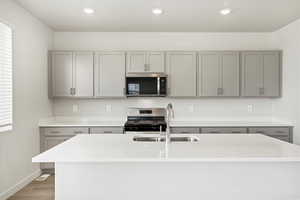 Kitchen featuring gray cabinetry, appliances with stainless steel finishes, an island with sink, light stone countertops, and light wood finished floors
