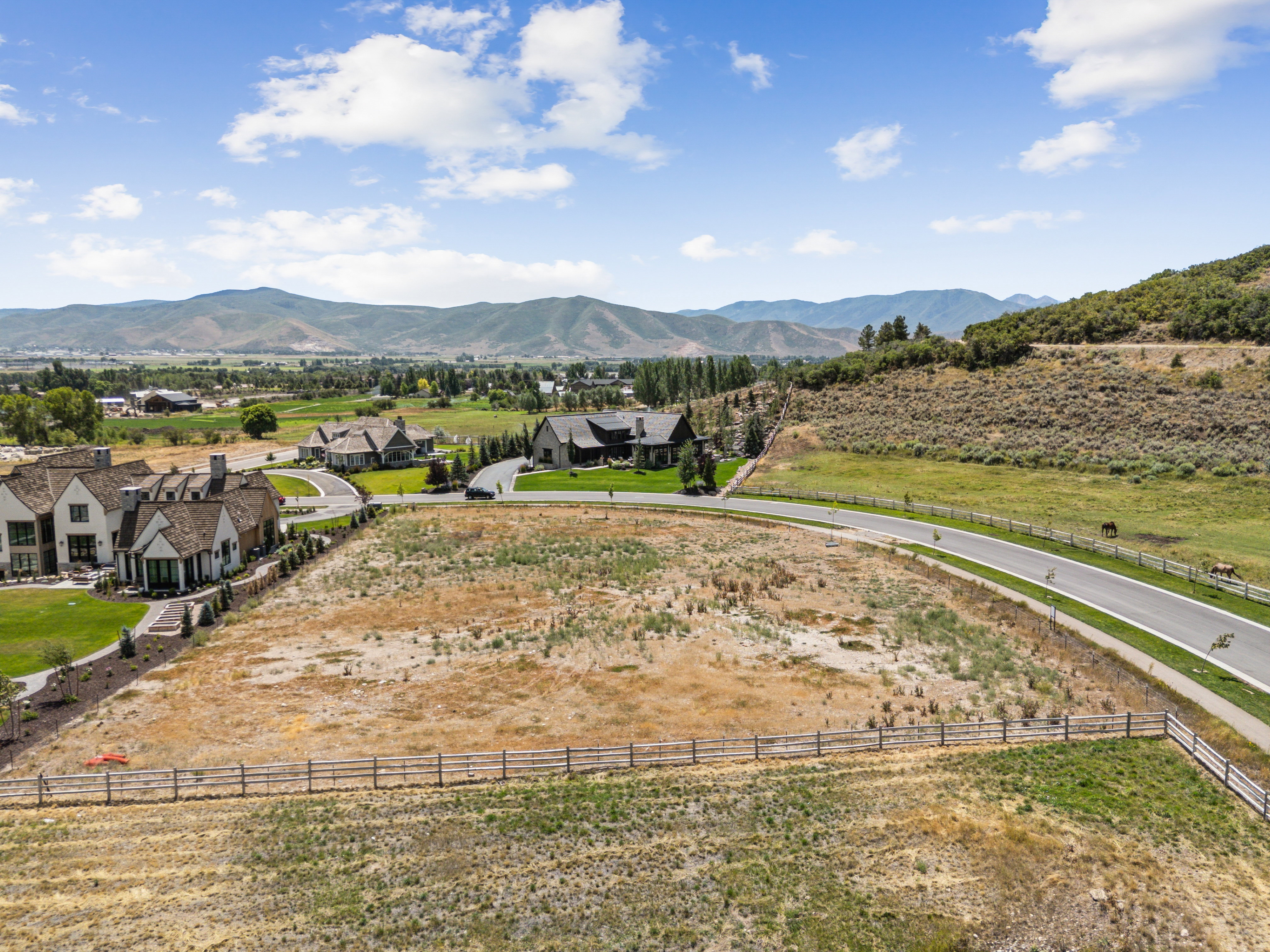 Expansive land parcel featuring distant mountain ranges, surrounding residential properties, paved road access, and a rustic wooden fence