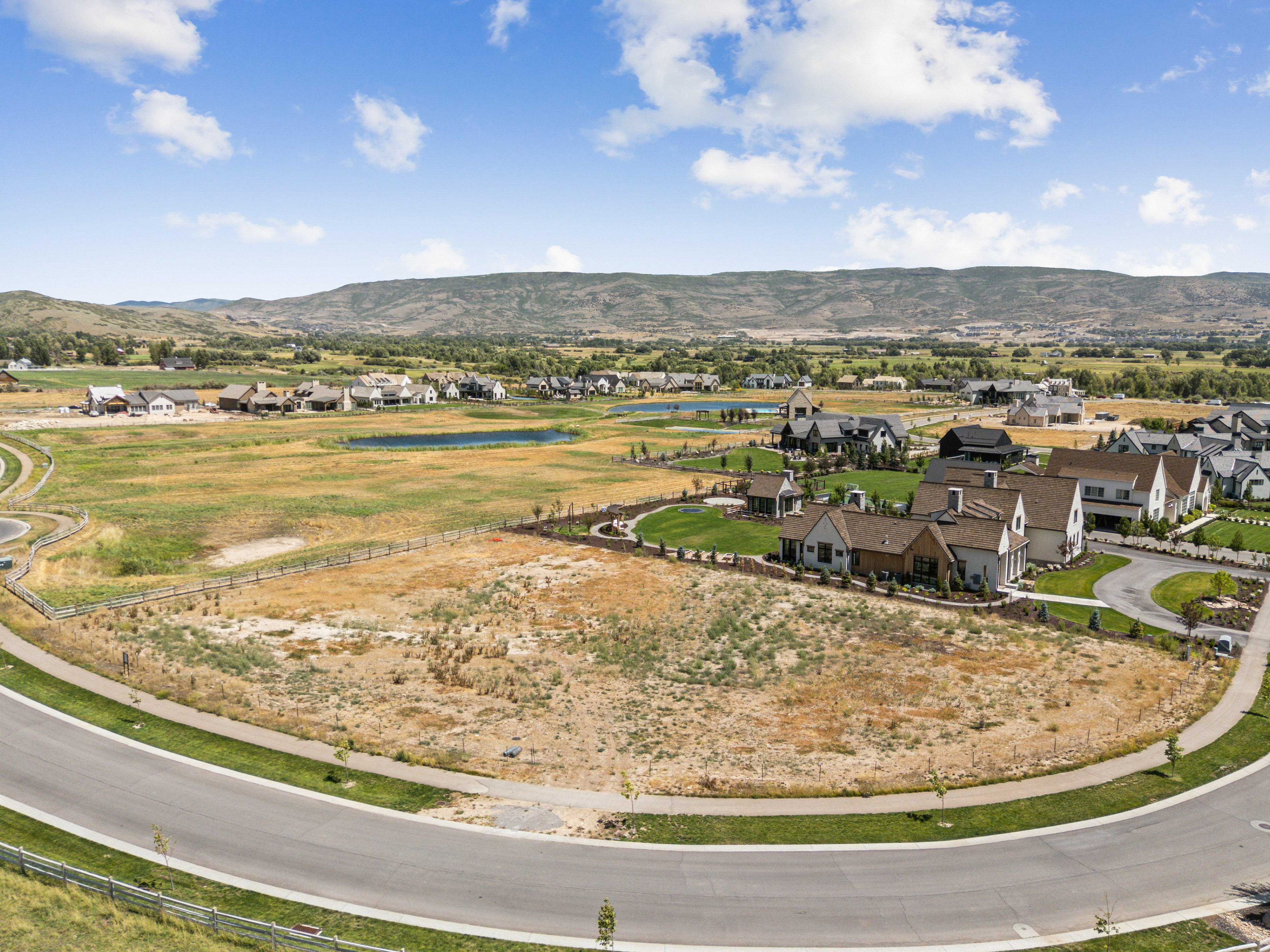 Expansive undeveloped land parcel featuring a paved road frontage, established residential properties nearby, and distant mountain scenery