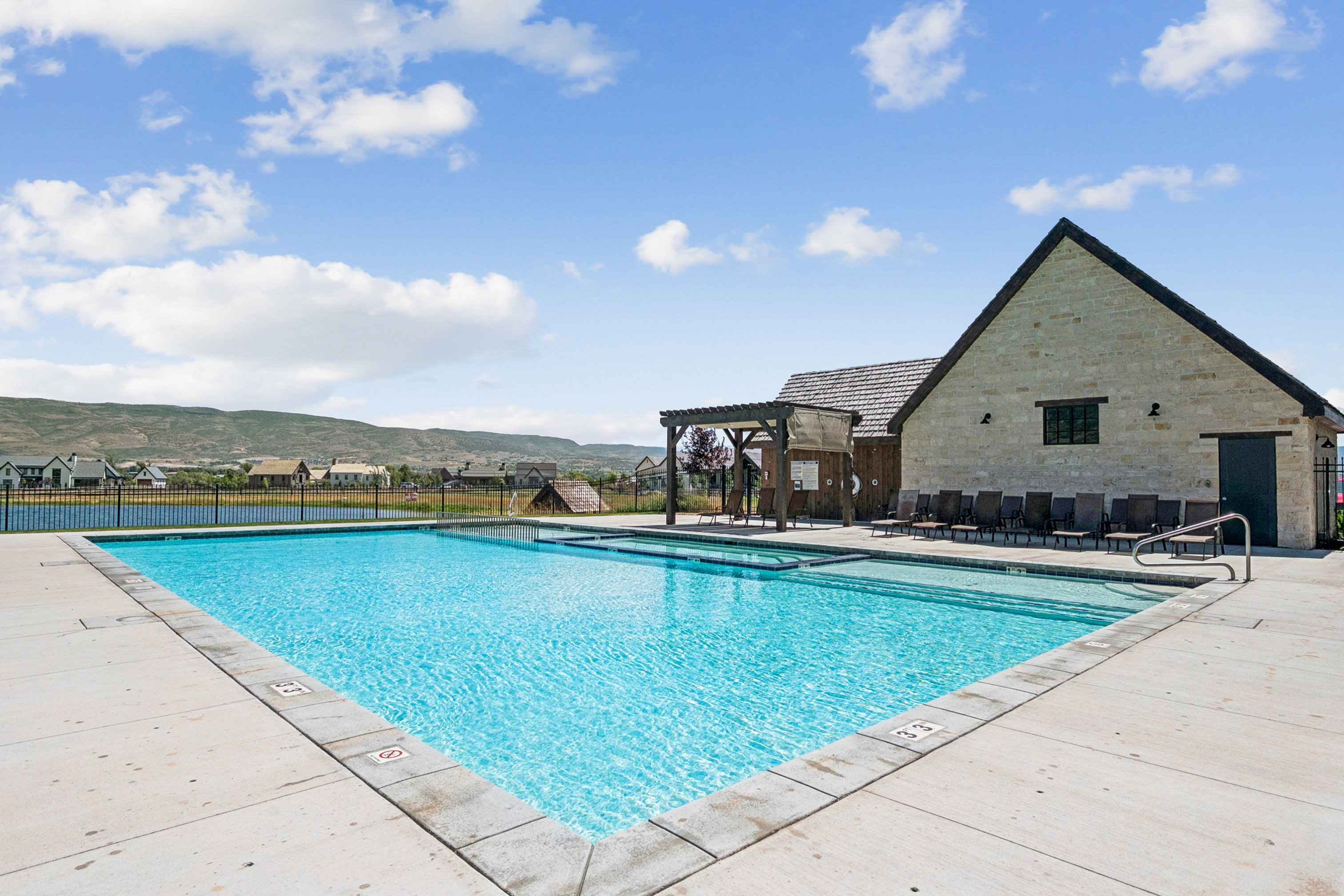 Outdoor swimming pool with concrete decking, a stone-clad building with a dark roof, and a wooden pergola structure