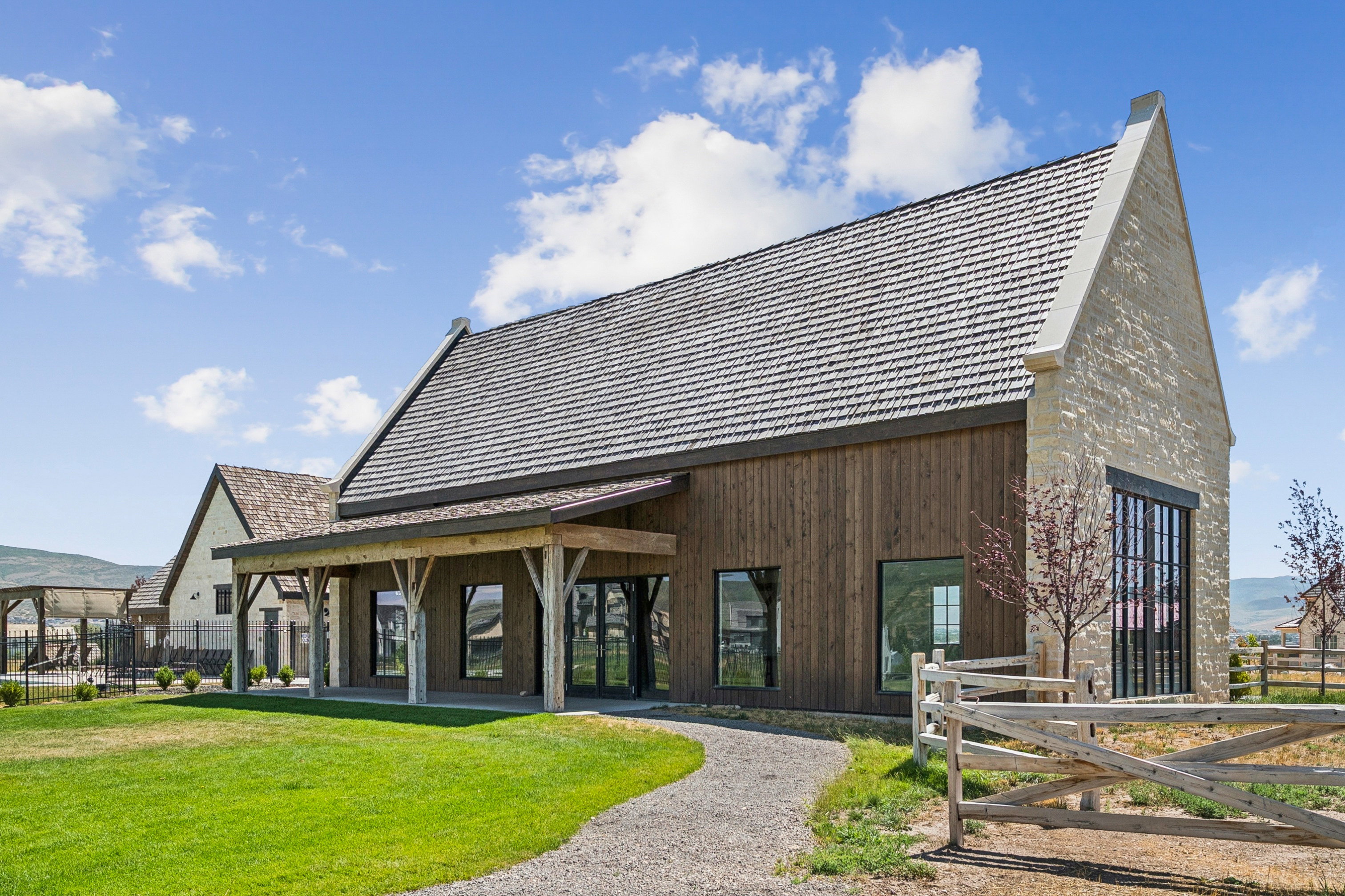 Modern farmhouse exterior with a prominent gravel pathway, featuring a covered porch with exposed timber beams, a wood-sided facade, and a stone gable end with a large multi-pane window