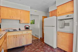 Kitchen featuring white appliances, light countertops, a textured ceiling, light brown cabinetry, and brick patterned floors