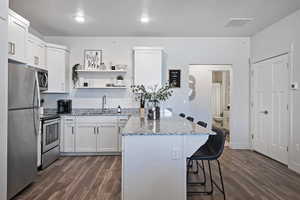 Kitchen with stainless steel appliances, a breakfast bar, dark wood-style flooring, light stone counters, and open shelves