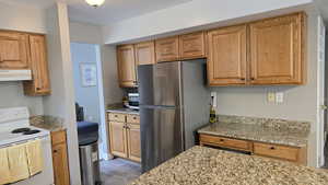 Kitchen with white electric stove, freestanding refrigerator, light wood finished floors, light stone counters, and under cabinet range hood